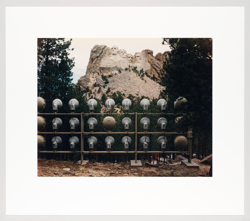 A photograph showing Mount Rushmore in the background with the carved faces of four U.S. presidents. In the foreground, there is a large array of spherical satellite dishes mounted on a grid structure. Trees flank the sides of the image, and the sky is overcast.