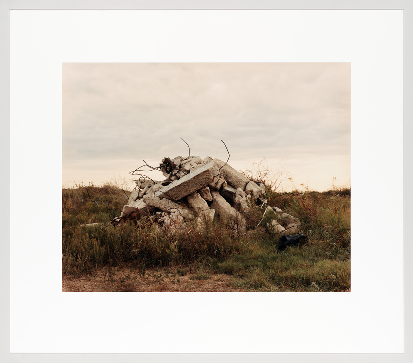 A pile of rubble consisting of broken concrete and twisted rebar sits in a grassy field under an overcast sky.