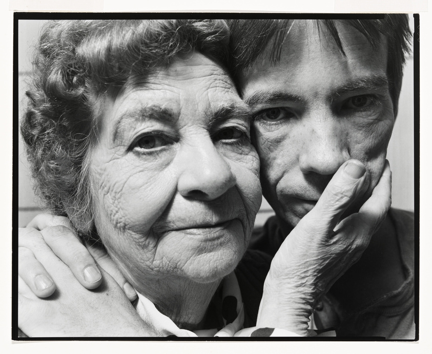 A black and white close-up photograph of an elderly woman and a younger person. The woman is smiling gently at the camera with her hand on the shoulder of the younger person, who is facing forward with a serious expression and their chin resting on the woman's hand.