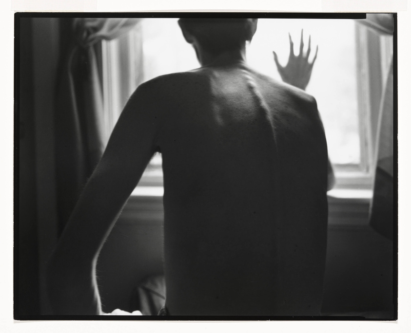 A black and white photograph capturing the back of a person standing in front of a window, with one hand raised against the glass pane. The person's back is bare, and the natural light from the window highlights the contours of their shoulder blades and arm.