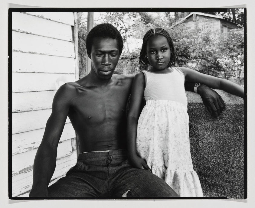 A black and white photograph featuring a shirtless man and a young girl sitting next to each other. The man, looking down, has his arm extended along the back of a bench, while the girl, wearing a white floral dress, stands with her arm resting on his, looking directly at the camera with a serious expression. They are outdoors, with trees and a white wooden structure in the background.