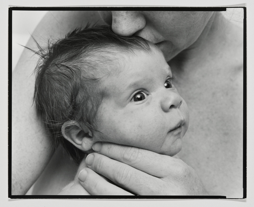 A black and white photograph capturing a tender moment between a baby and an adult. The baby, with wide eyes and wispy hair, is being cradled and gently held on the cheek by the hand of an adult, whose chin rests atop the baby's head. The baby's gaze is directed off-camera, showing a sense of curiosity or wonder. The close-up shot emphasizes the intimate and protective gesture.