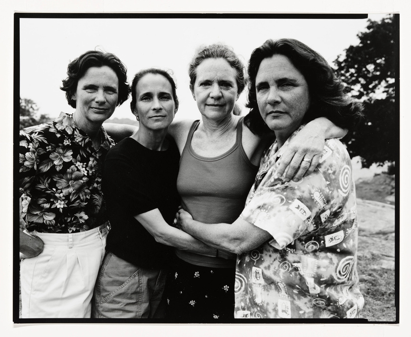Four women stand closely together, posing for a black and white photograph. They are dressed in casual clothing, with two wearing floral patterns, one in a plain black top, and the other in a tank top. The background is out of focus, suggesting an outdoor setting. Their expressions are serious and they appear to be comfortable with each other, possibly indicating a close relationship.