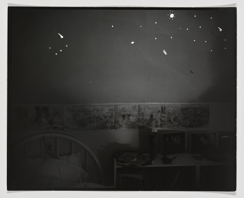 A monochrome photograph of a bedroom with glow-in-the-dark star stickers on the ceiling. Below, there's a bed with a person lying down, partially covered by bedding. A wall-mounted series of drawings or prints and a shelf with books, framed photographs, and a small TV are visible. The room has a cozy, lived-in atmosphere.