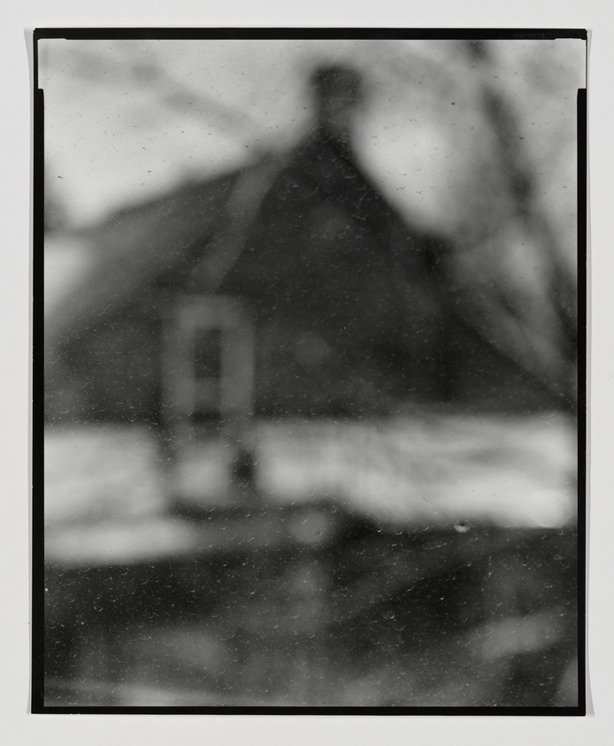 A monochrome, blurry photograph of a house with a prominent gable and a window visible on the front facade. The image has a vintage or aged appearance with dust and scratches on the surface, and the edges of the photo are bordered with black, suggesting it may be an old film photograph. The background is indistinct, possibly showing trees or other vegetation.