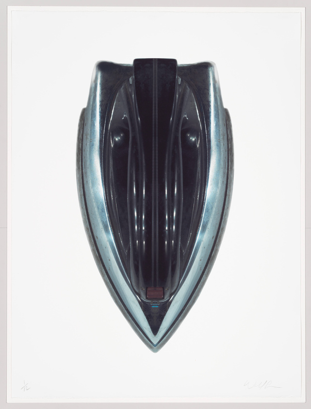 A glossy, metallic sculpture resembling a stylized mask or face, with a symmetrical design featuring a central vertical ridge flanked by curved, rib-like structures. The piece has a pointed chin and a broad, shield-like forehead, presented against a white background with artist's signatures at the bottom corners.