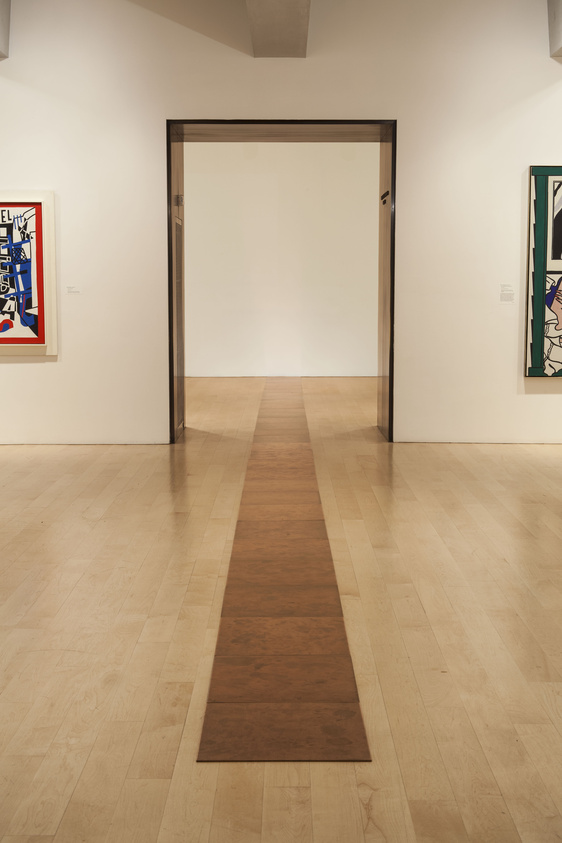 A minimalist art gallery interior with a wooden floor and framed artworks on white walls, viewed through an open doorway.