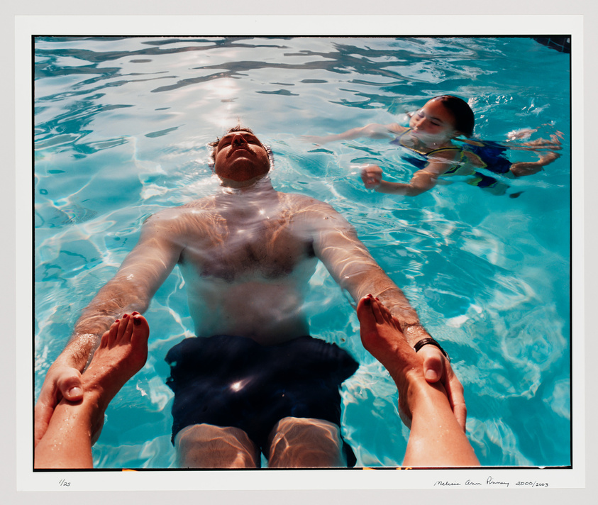 A man is floating on his back in a clear blue swimming pool, his arms extended towards the camera with a child swimming in the background to his right. The sun illuminates the water, creating a tranquil scene. The image is framed with a white border that includes a signature and edition number in the lower left corner and a name and date in the lower right corner.