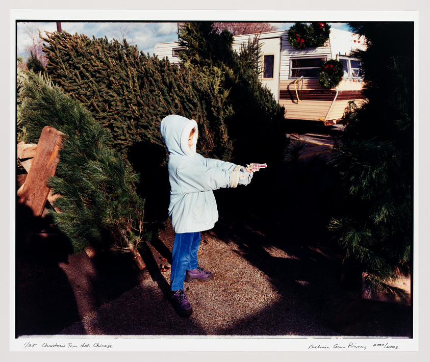 A child in a blue hooded jacket and jeans is pointing a toy gun to the right side of the frame, standing in a lot with Christmas trees and a decorated trailer in the background. The image has handwritten inscriptions at the bottom indicating it's a photograph of a Christmas tree lot in Chicago by Melissa Ann Pinney, dated April 2003.
