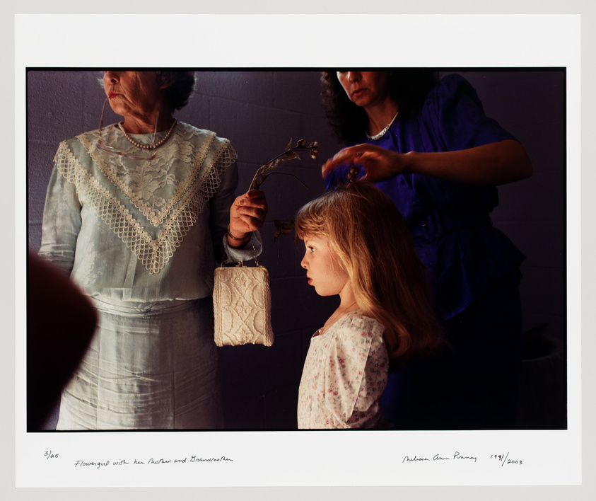 A photograph capturing three generations of women, with a young girl in the foreground looking to the side, an older woman holding a purse and a branch in the middle, and another woman in the background adjusting the young girl's hair. The image has a handwritten caption at the bottom indicating the title and the artist's signature with a date.