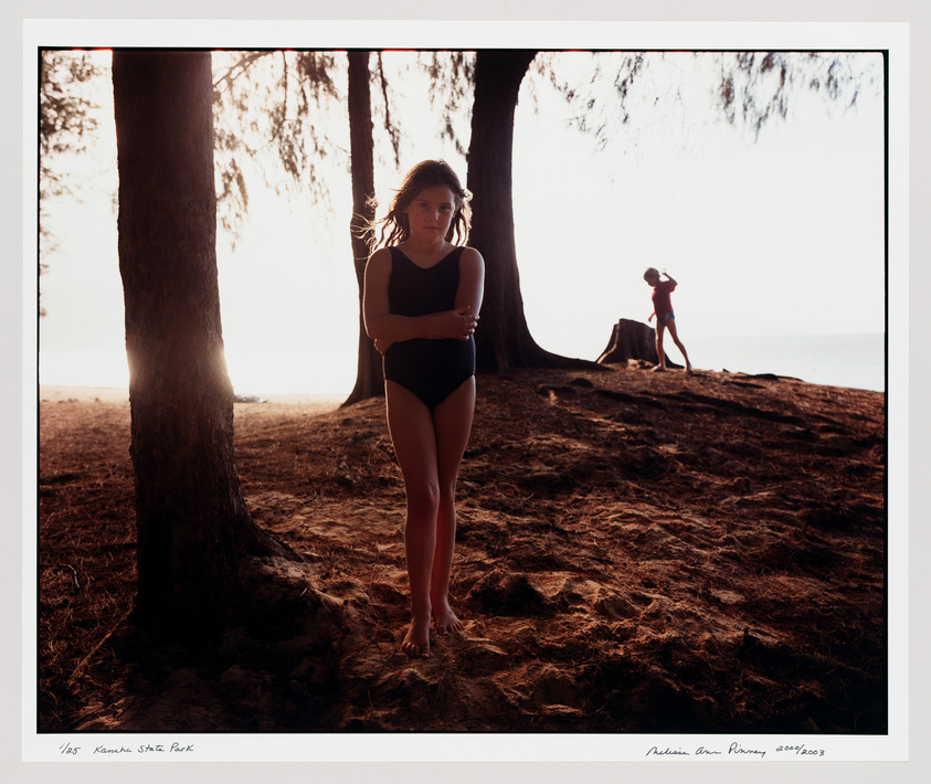 A photograph capturing two individuals at a beach during sunset or sunrise. The foreground shows a young woman in a black swimsuit standing near a tree, looking pensively at the camera with her arms crossed. In the background, another person is seen in a playful pose atop a tree stump, silhouetted against the bright sky. The scene is framed by trees and the ground is covered with textured sand. The lighting creates a warm, golden hue throughout the image.