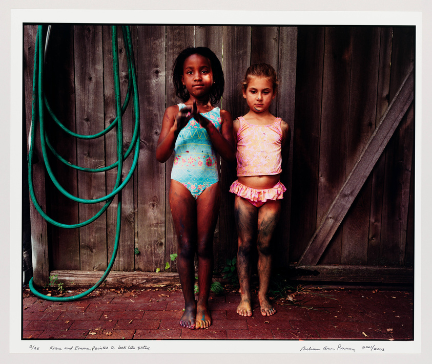 Two young girls stand side by side against a wooden fence, one wearing a blue swimsuit and the other in a pink tankini with a ruffled skirt. A green garden hose is coiled on the left side of the image. The girls have dirt on their legs, suggesting outdoor play, and they look directly at the camera with neutral expressions. The photograph has a caption at the bottom indicating the names of the subjects and the artist's signature with a date.