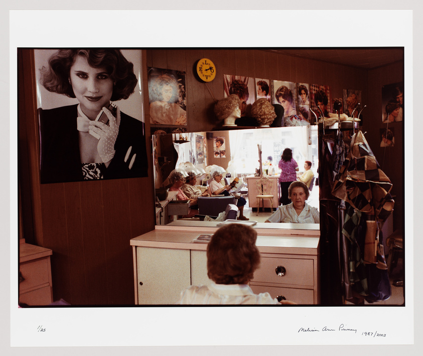 A photograph of the interior of a bustling beauty salon with multiple mirrors reflecting the image of a woman sitting at a styling station. The walls are adorned with various portraits of women with different hairstyles. A large poster of a woman with a glamorous look dominates the left side of the image, while the salon's activity is visible in the background. A clock and some hair wigs can also be seen on the wall. The scene captures the essence of a typical hair salon from a bygone era.