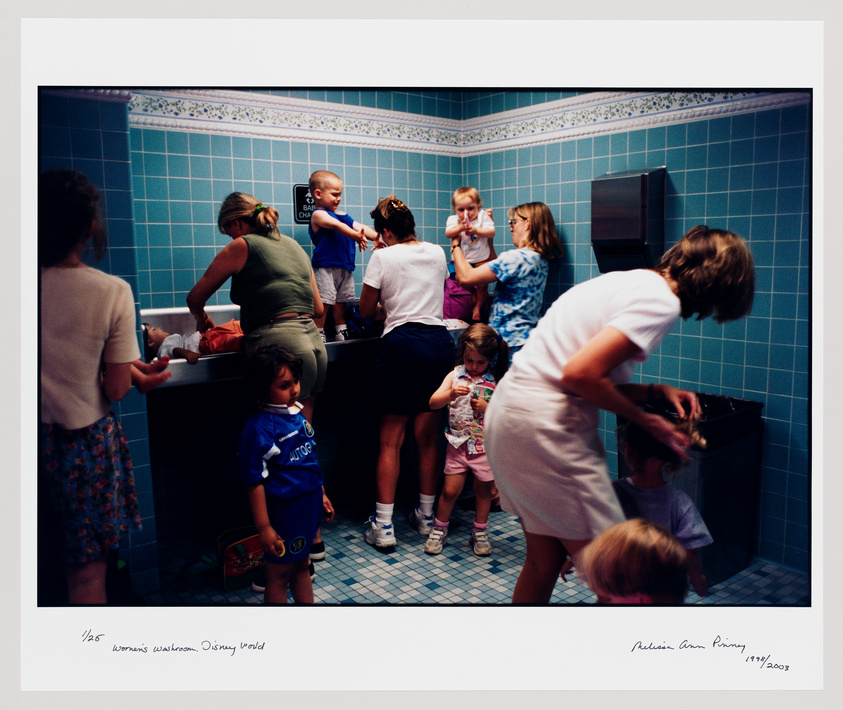 A busy scene inside a women's restroom with multiple people. Several women and children are engaged in various activities such as washing hands, looking in the mirror, and tending to children. The room is tiled in blue with a large mirror reflecting the activity. There's a handwritten caption at the bottom indicating the location and date.