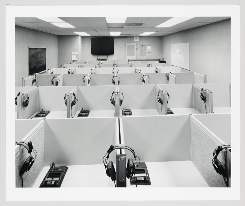 A monochrome photo of an empty call center with rows of cubicles, each equipped with a headset and a computer, under fluorescent lighting. A large screen hangs on the front wall, and various notices are pinned to a bulletin board on the back wall.