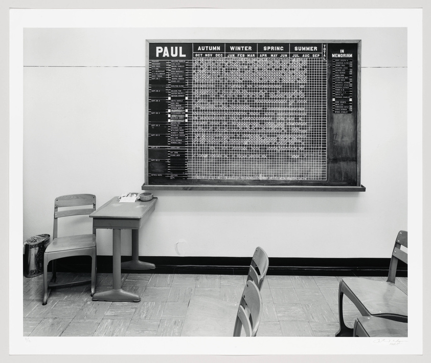 A black and white photograph of an interior room with a large scheduling board labeled "PAUL" on the wall. The board is divided into sections for Autumn, Winter, Spring, and Summer, with a grid of names and dates. To the left, there's a small desk with papers and a telephone, and in the foreground, several chairs are arranged facing the board. The room appears to be an office or waiting area.