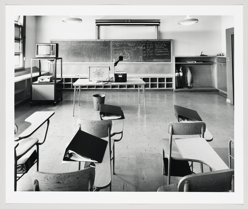 A black and white photograph of an empty classroom with individual student desks facing a chalkboard filled with writing. On the left, there is a TV on a cart and a projector on a table. The room has a retro feel, suggesting the photo may be from the mid to late 20th century.