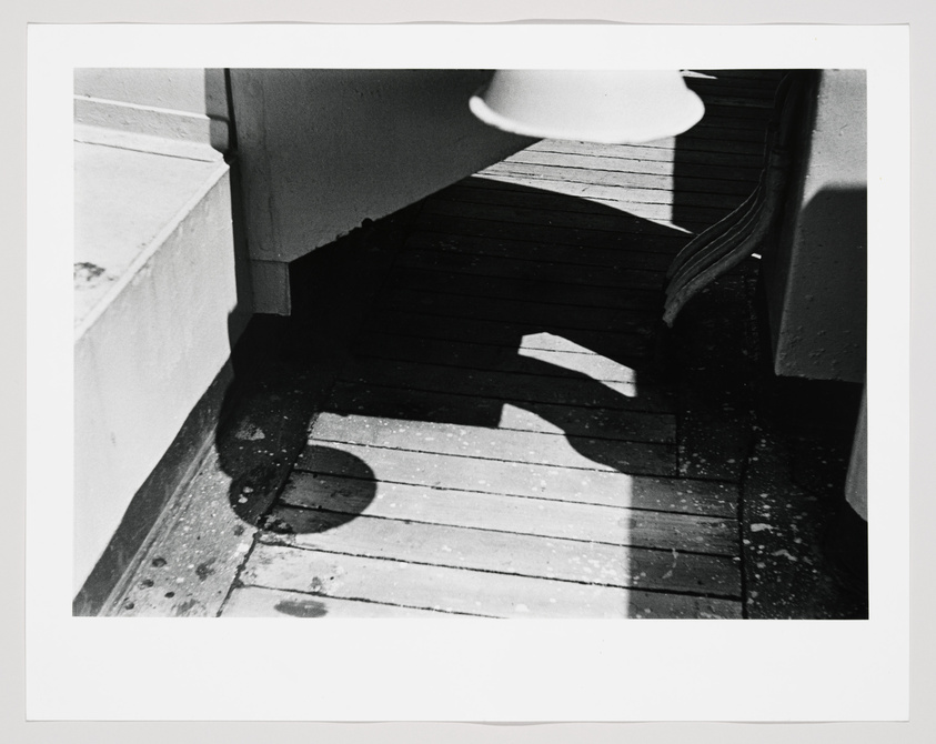 Black and white photograph capturing the shadow of a person and a lamp post on a wooden deck, with the shadow suggesting the person is wearing a hat.