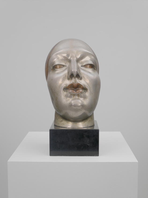 A metallic sculpture of a human head with a serene or expressionless face, displayed on a black pedestal atop a white plinth against a neutral background.