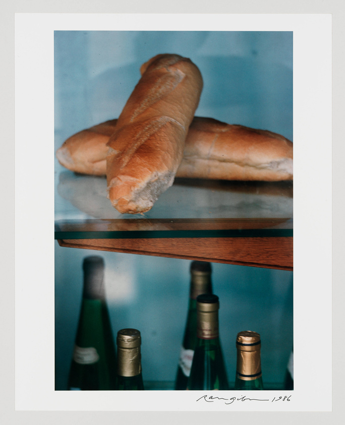 A photograph of a baguette placed on a glass shelf, with the reflection of the bread visible on the surface. Below the shelf, the tops of several wine bottles can be seen, suggesting the setting may be a kitchen or a dining area. The image includes a signature and the year 1986 at the bottom right corner.