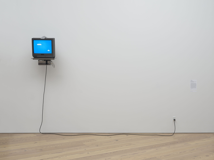 A CRT monitor displaying a blue screen with clouds mounted on a white wall above a DVD player, connected to a power outlet by a long cable.