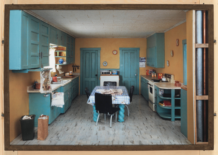 A cozy vintage kitchen with teal blue cabinets and doors against mustard yellow walls. The room features a checkered blue and white tablecloth on a dining table, various kitchen utensils, and a mix of decorative and practical items on the countertops. A calendar hangs on the wall, and there is a glimpse of a white tiled backsplash. The floor is covered with light-colored tiles, and there are two trash bins near the entrance. The image is framed as if looking through a window or doorway.