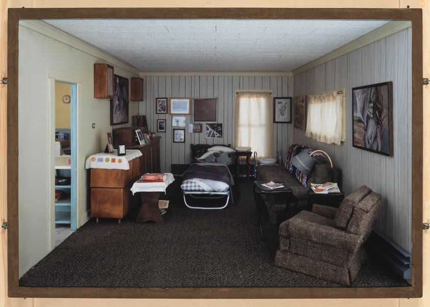 A cozy, vintage living room with wood-paneled walls and a shaggy brown carpet. The room features a single bed with a plaid blanket, a wooden dresser with a mirror and personal photos, and a small wooden nightstand with a lamp. A recliner and a sofa with patterned throws are positioned near a coffee table with magazines. The walls are adorned with various framed pictures and paintings, and two curtained windows allow soft light to filter in. The view is from the doorway, giving a sense of stepping into someone's personal space.