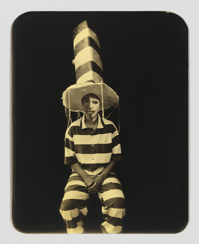 A sepia-toned photograph of a person wearing a vertically striped hat and matching shirt and pants, sitting with hands clasped between knees. The outfit resembles a classic prisoner uniform, and the hat is exaggeratedly tall. The person's face is painted white with dark circles around the eyes, and they are looking directly at the camera with a neutral expression. The background is plain and dark, highlighting the subject.