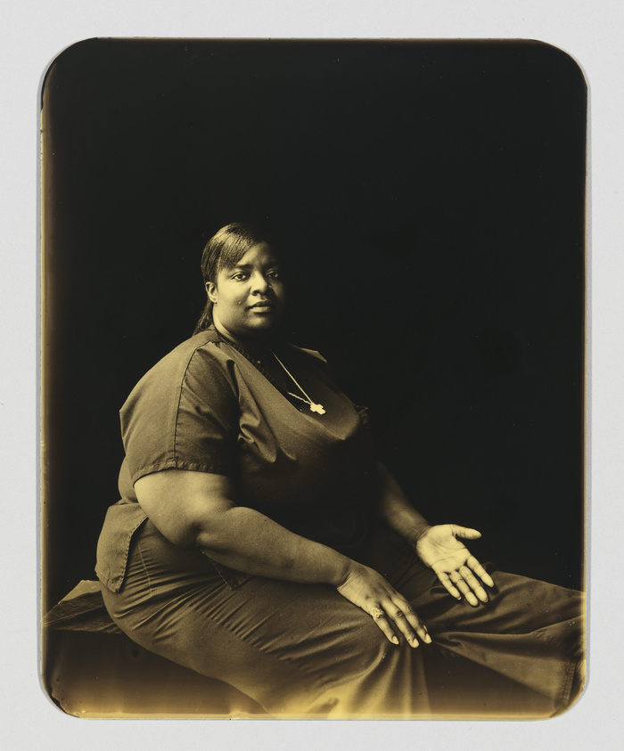 A sepia-toned photograph of a seated woman with a calm expression, wearing a brown dress and a necklace with a cross pendant. She is positioned slightly to the side, with her hands resting on her lap against a dark background.