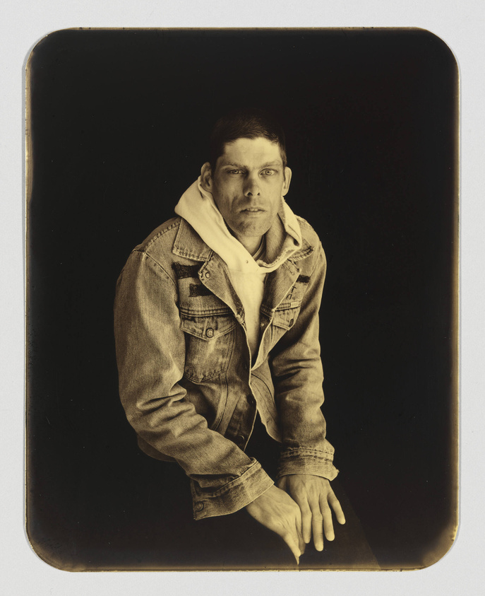 A sepia-toned photograph of a man seated with a serious expression, wearing a denim jacket over a hoodie, against a dark background.