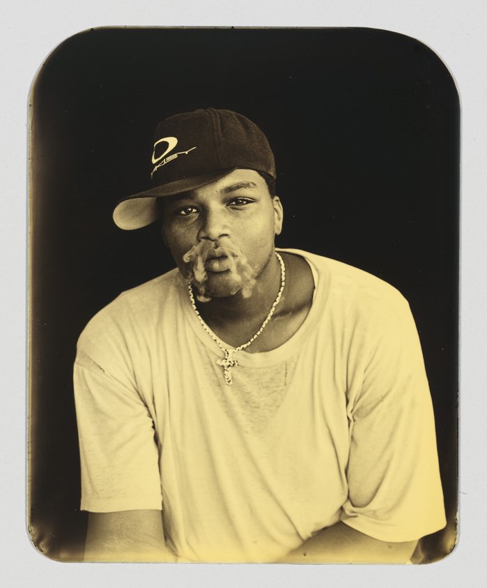 A sepia-toned portrait of a young man wearing a baseball cap and a casual t-shirt, exhaling smoke with a relaxed expression. He has a chain necklace with a pendant, and the image has rounded corners, giving it a vintage appearance.