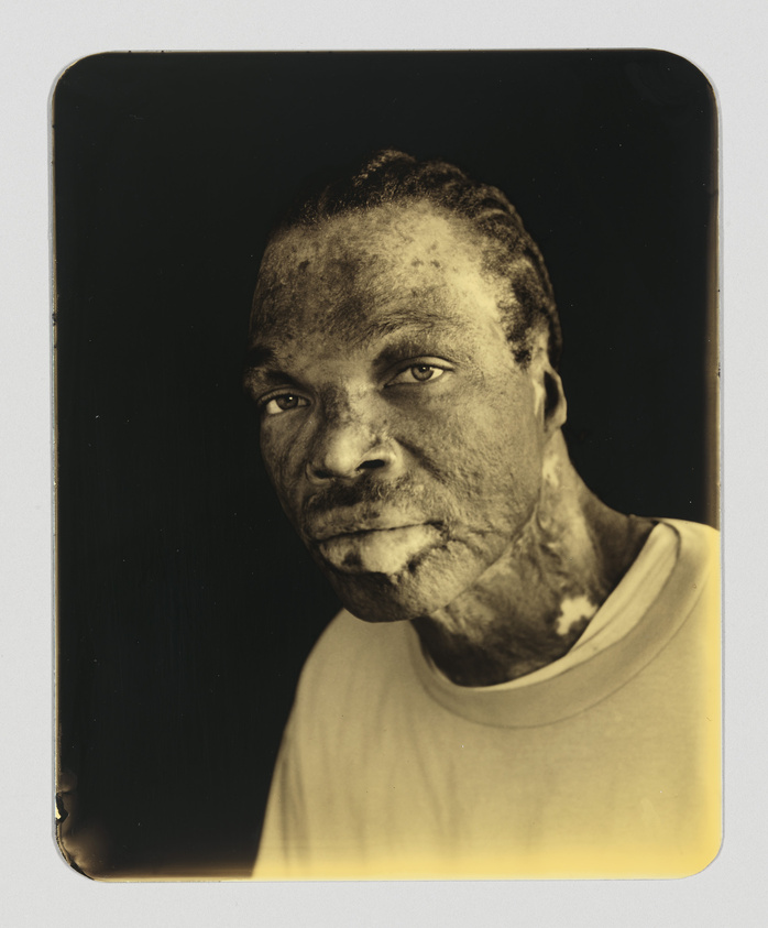 A sepia-toned portrait of a man with a contemplative expression, looking directly at the camera. The image has a vintage appearance with rounded corners and a black background, highlighting the details of the man's face and his light-colored T-shirt.
