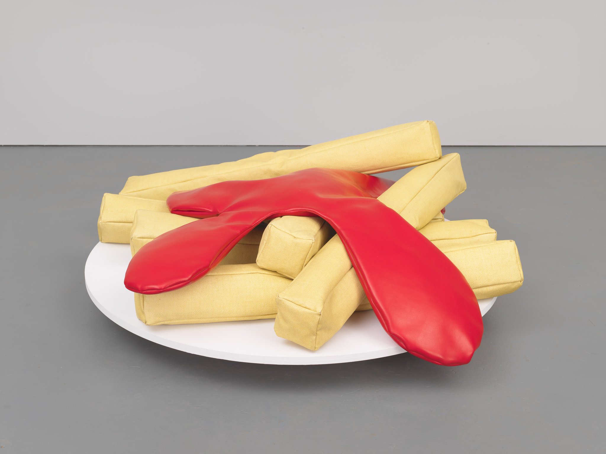 A sculpture resembling a pile of oversized, realistic-looking French fries in a yellow color, with a large red ketchup-like form draped over them, all presented on a round white platter against a grey background.