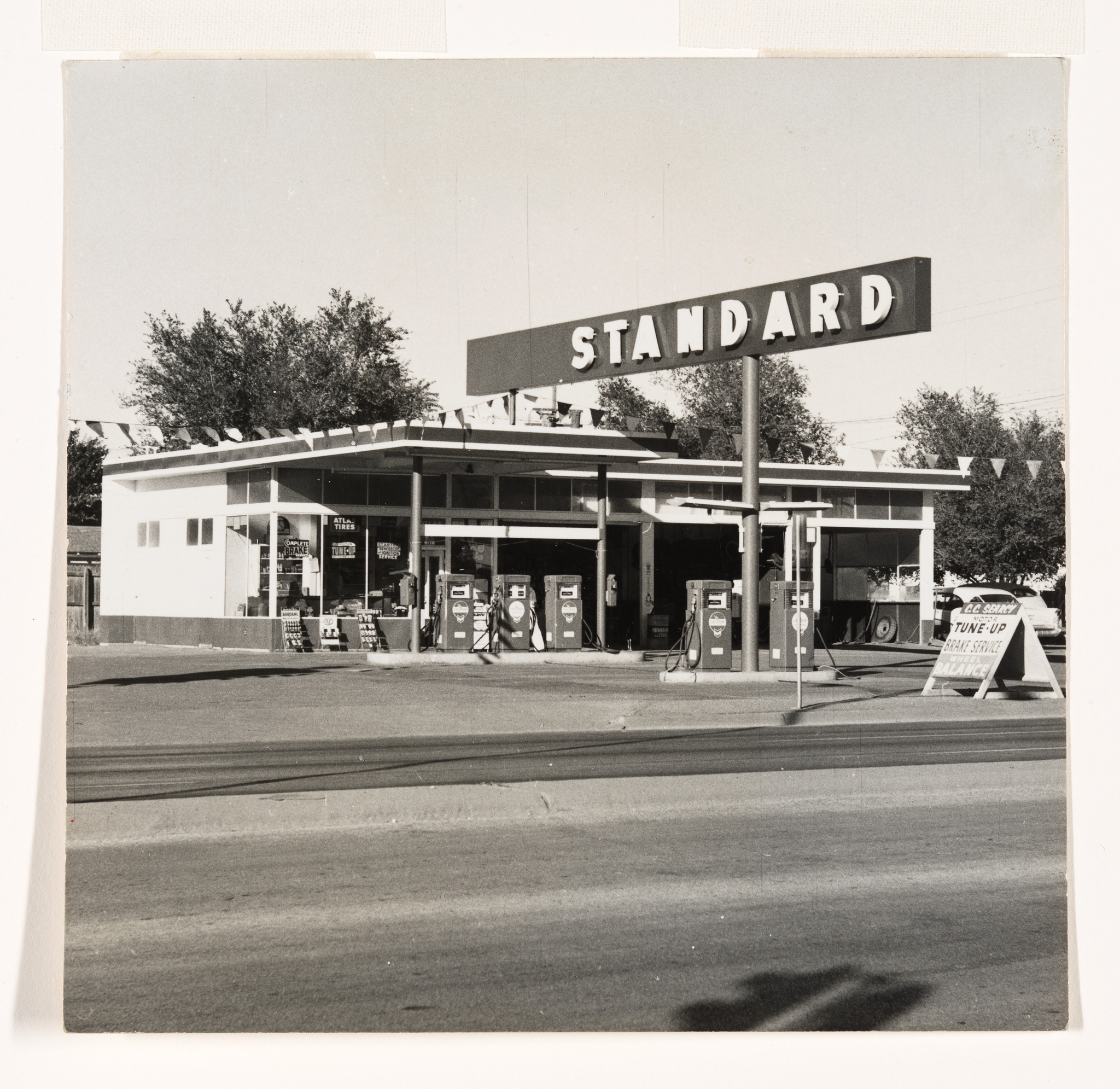 A vintage black and white photo of a Standard gas station with multiple fuel pumps and a large sign with the company name. The station has a mid-20th-century architectural style, and there are promotional banners and a sign advertising a tune-up service. The street in the foreground is empty, and there are no visible people.