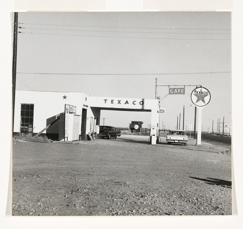 A vintage black and white photo of a Texaco gas station with a cafe sign, featuring a classic car at the pump and another car parked nearby. The station has a large Texaco star logo and the scene has a mid-20th-century Americana vibe.