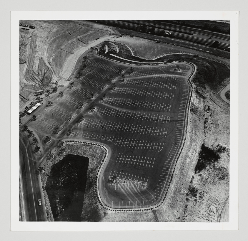 Aerial black and white photograph of a large, empty parking lot with organized rows and curved ends, surrounded by undeveloped land with visible tire tracks and a few scattered vehicles on the periphery.