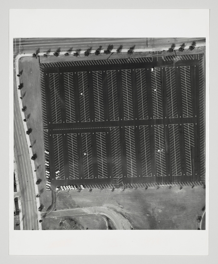 Aerial black and white photograph of a large parking lot with numerous empty spaces, bordered by a road with a few vehicles and a line of trees. The parking lot shows geometric patterns of parked cars and empty rows, with surrounding areas of undeveloped land.