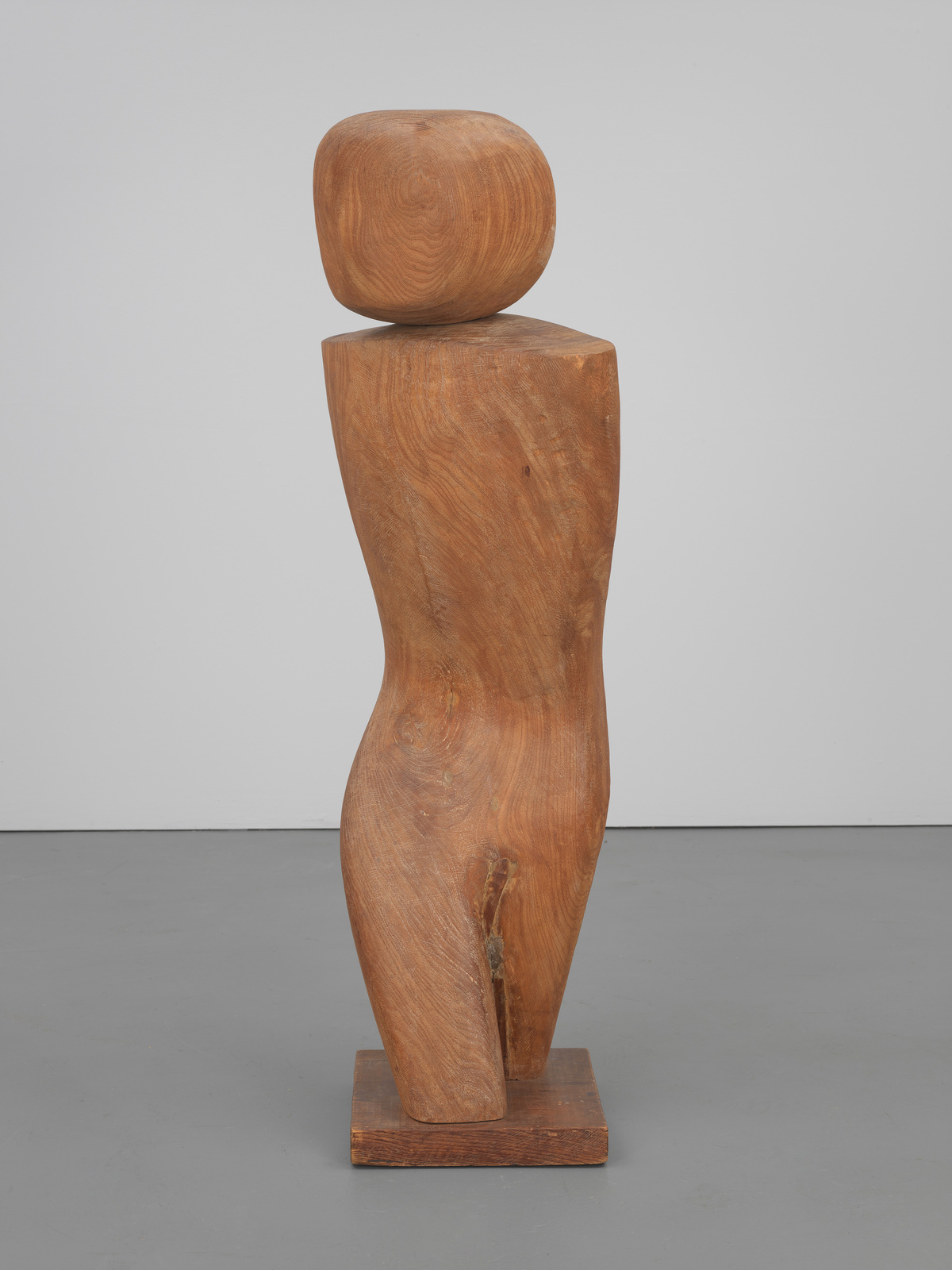 A wooden sculpture displayed against a white background, featuring an abstract human form with a rounded head atop a tapered, columnar body, all with a smooth, polished surface and the natural grain of the wood visible. The sculpture is mounted on a flat square base.