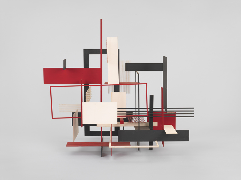 A modern abstract sculpture featuring geometric shapes in red, black, and white, with linear elements and planes intersecting at various angles against a neutral background.