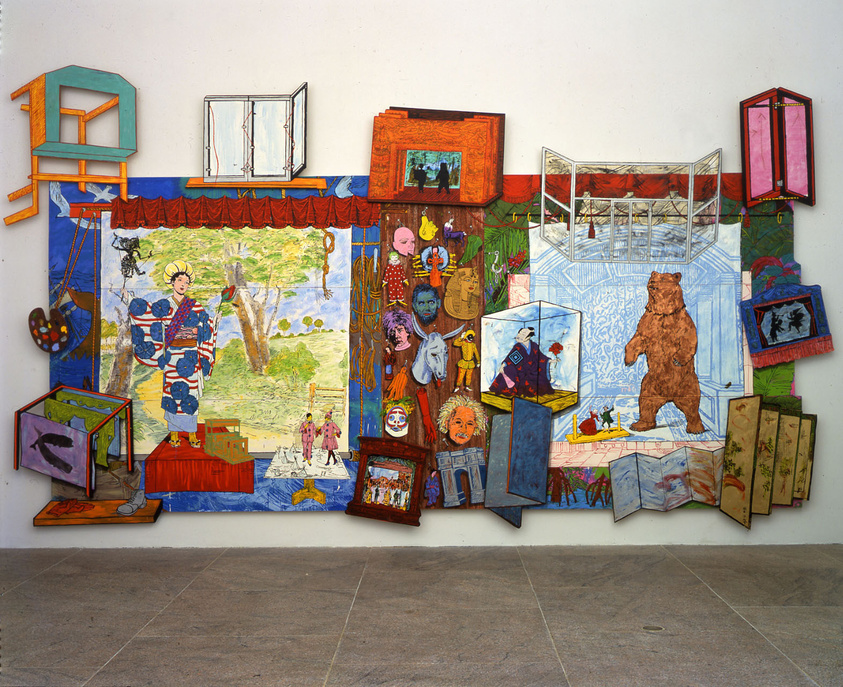 A colorful and eclectic mixed-media art installation displayed on a white wall, featuring a central tapestry with a pastoral scene and various figures, flanked by an assortment of painted frames, boxes, and sculptural elements that extend into the third dimension. Some frames contain images of people and animals, while others are open, revealing the wall behind. The artwork has a playful and whimsical quality, with a bear figure standing in the center and a variety of textures and patterns throughout.