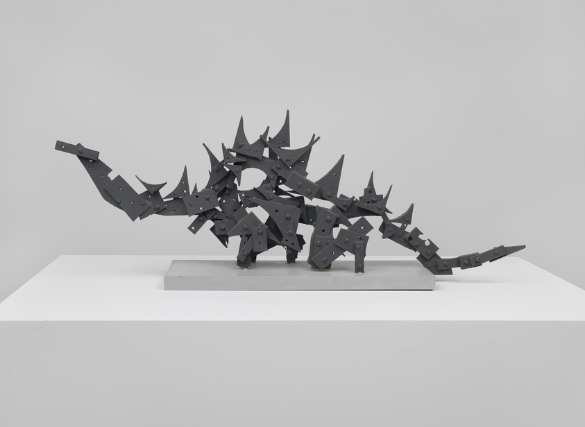 A modern, abstract metal sculpture resembling a stylized creature with numerous spikes and geometric shapes, displayed on a white pedestal against a grey background.