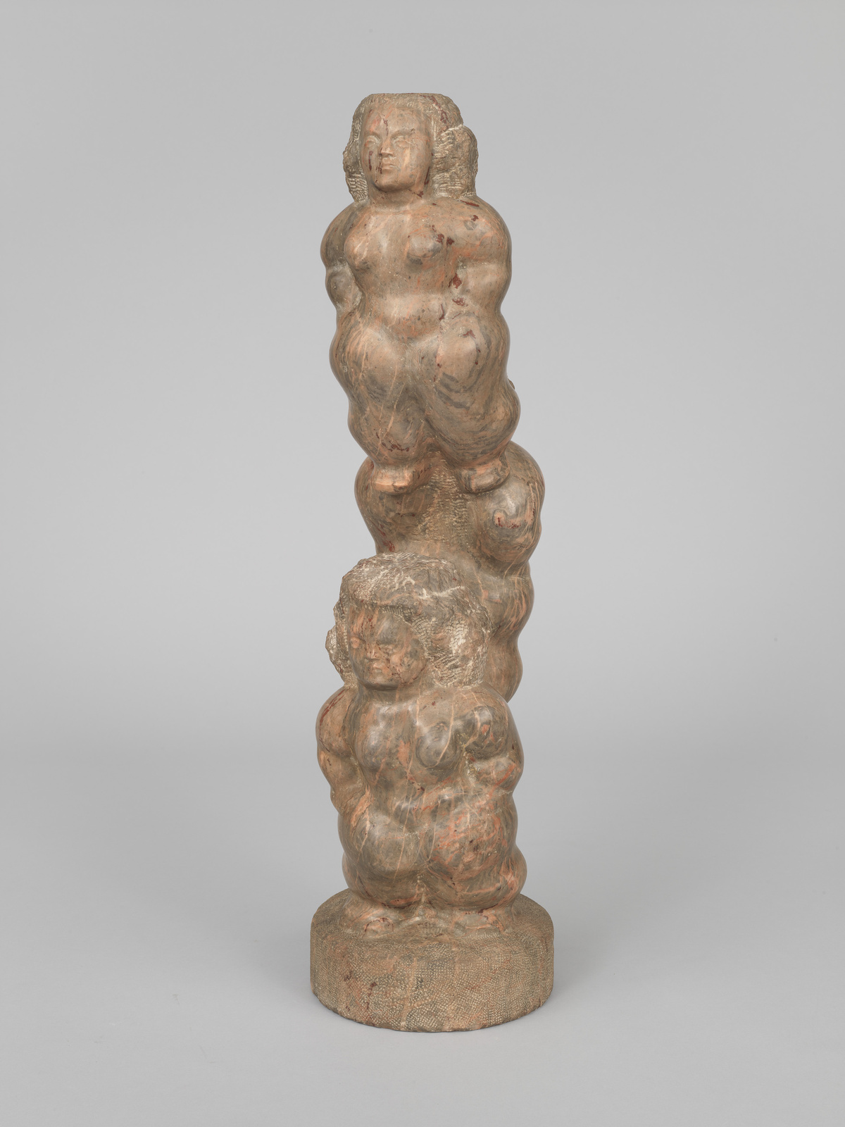 A carved wooden sculpture depicting a vertical stack of three rounded, abstract human figures with discernible heads, torsos, and limbs, set against a neutral background. The figures appear to be fused together, creating a totem-like column on a circular base. The wood has a worn, patinated surface with visible grain and remnants of paint or finish.