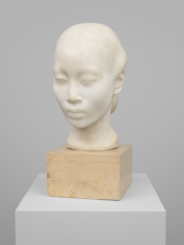 A marble sculpture of a human head with a serene expression, displayed on a square stone pedestal atop a white plinth against a grey background.