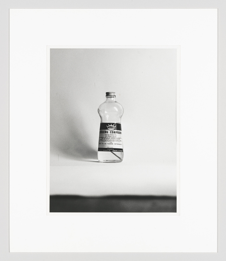 A black and white photograph of an old-fashioned glass bottle labeled "Rubbing Compound" centered on a plain background, framed with a wide white border.