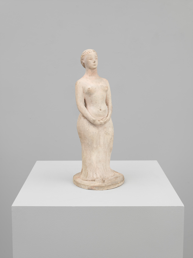 A white, ancient Greek-style statue of a standing female figure displayed on a white pedestal against a grey background. The statue appears to be made of marble or a similar material and shows signs of age and wear. The figure is depicted with her hands resting gently on her abdomen, her hair pulled back, and her gaze slightly downward.