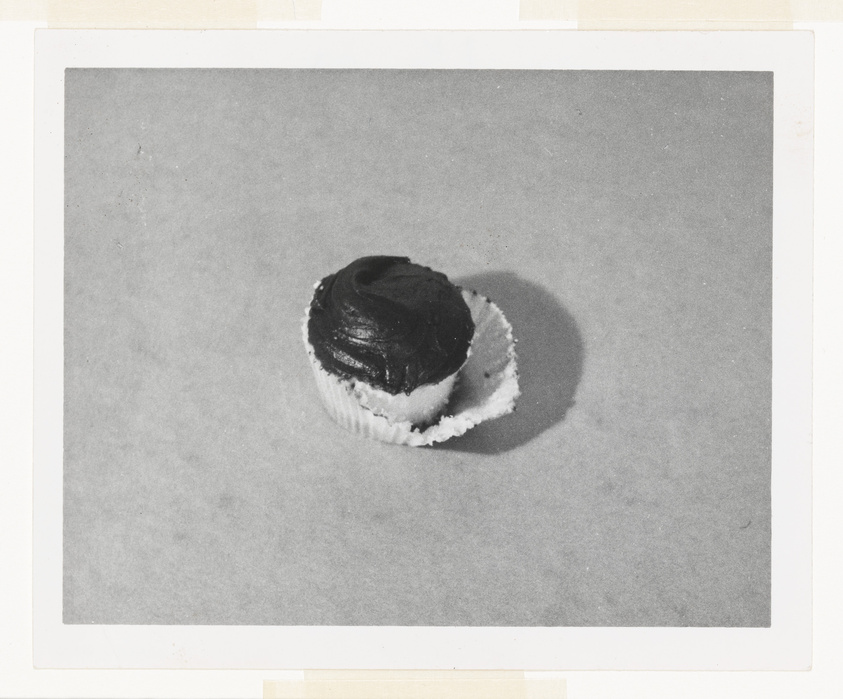 A black and white photo of a single cupcake with dark frosting on a plain background, mounted on a white border.