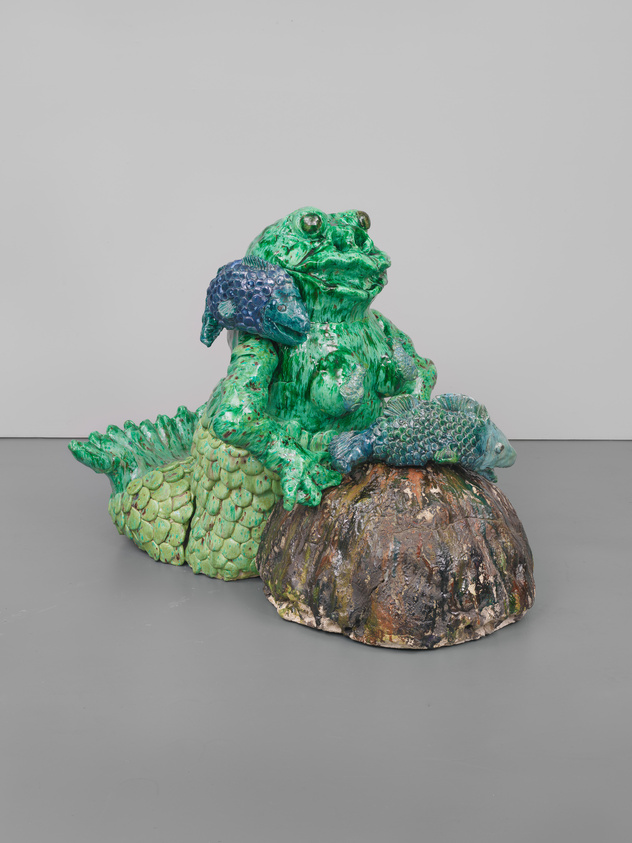 A colorful ceramic sculpture of a frog sitting on a stump, with a textured and glossy finish, predominantly in shades of green with some blue accents. The frog has a whimsical expression and is depicted in a seated position with its hind legs visible and front limbs resting on its body. The sculpture is set against a plain, light-colored background with a gray floor.