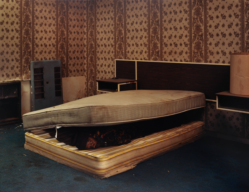 A person is hiding under a mattress in a room with vintage wallpaper and a blue carpet. The room appears old and neglected, with a fireplace and a wooden headboard visible. The mattress is lifted at an angle, revealing the person underneath.