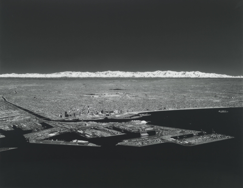 A black and white aerial photograph showcasing a dense urban cityscape with numerous buildings, adjacent to a large body of water with docks and ships. In the background, a prominent mountain range capped with snow stretches across the horizon.