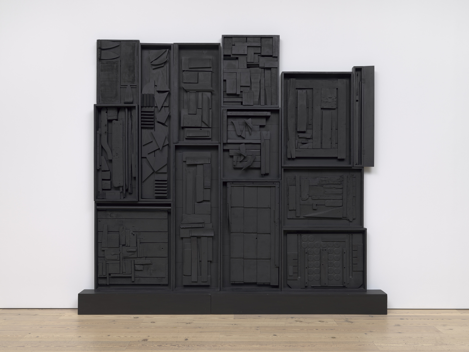 Black abstract wall sculpture with various geometric shapes and textures, displayed on a wooden floor.